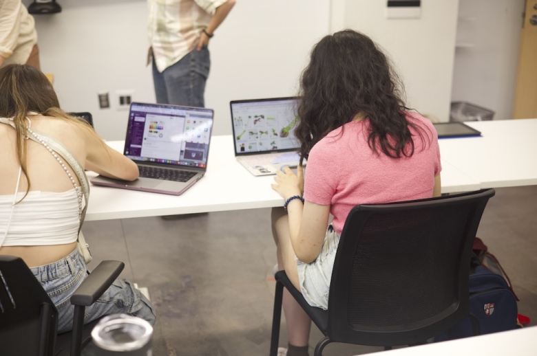 Photo taken behind two Tisch Summer High School Game Design students seated and facing toward their laptop computers